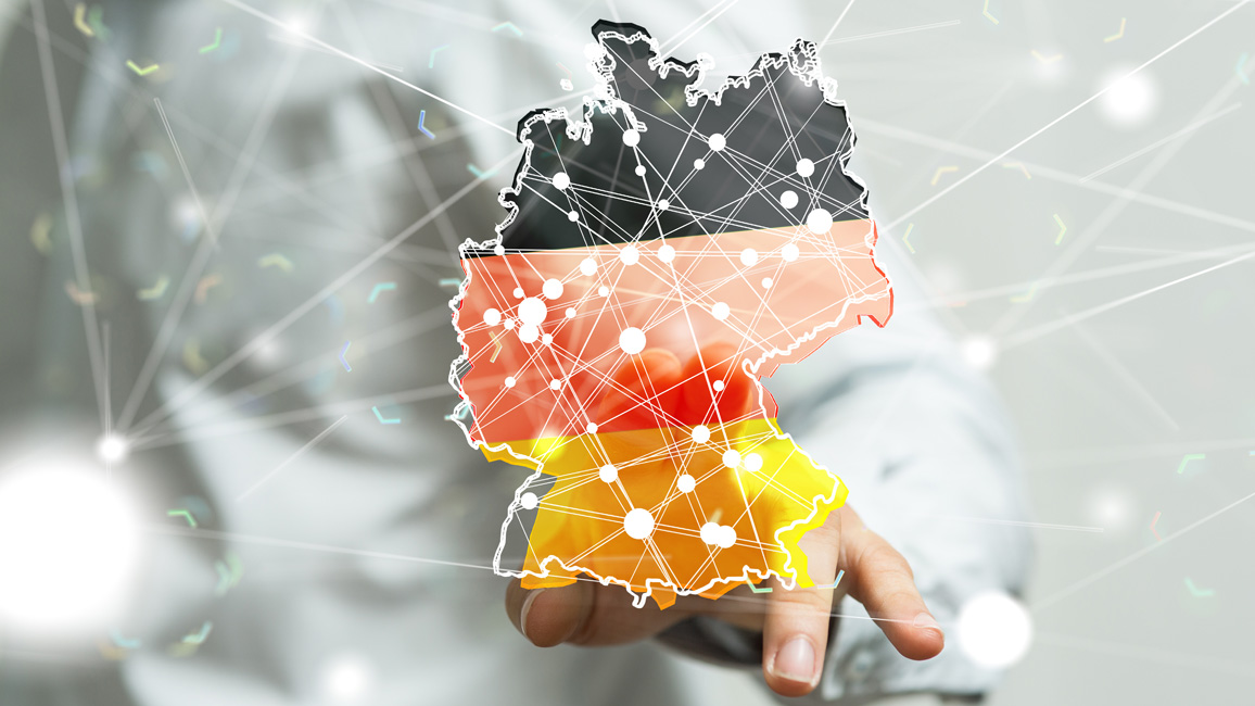 Outline of a map of Germany which is pigmented in the colours of the German flag: black, red and yellow. On top of it, there are white dots of different sizes connected by several white lines to a kind of network. In the blurred background, there is a person pointing at this figure.