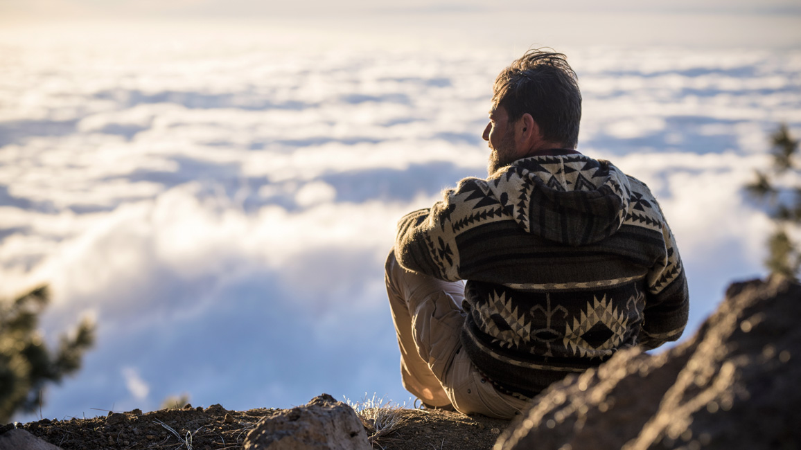 A man is sitting on the top of a mountain looking into the distance.