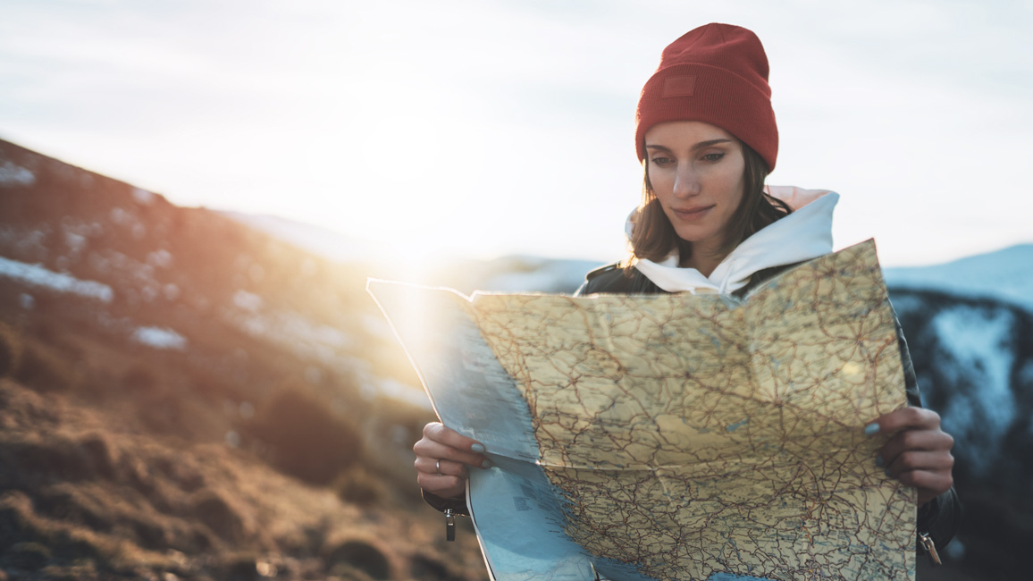 A female hiker with brown hair and a red hat looks concentrated on a map. In the background there is a blurred mountaineous landscape.