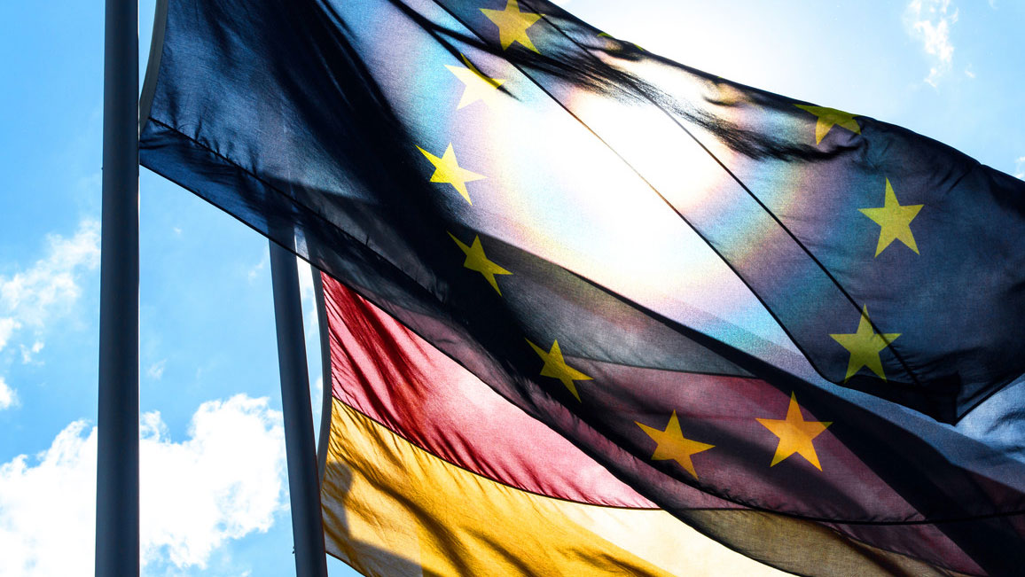 The EU flag is fluttering in the wind in front of the flag of Germany. In the background there are a few clouds in the sky and the sun is shining.