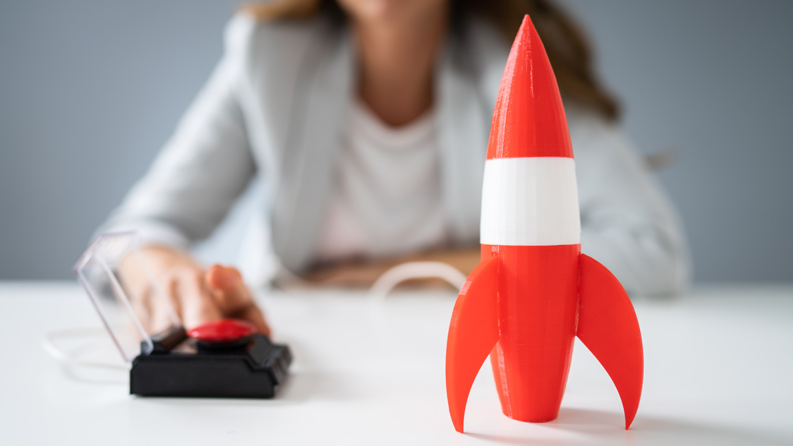 Eine rot-weiße Modell-Rakete steht auf einem Tisch. Im Hintergrund ist verschwommen zu sehen, dass eine Frau kurz davor ist, den dazugehörigen Start-Knopf zu drücken.