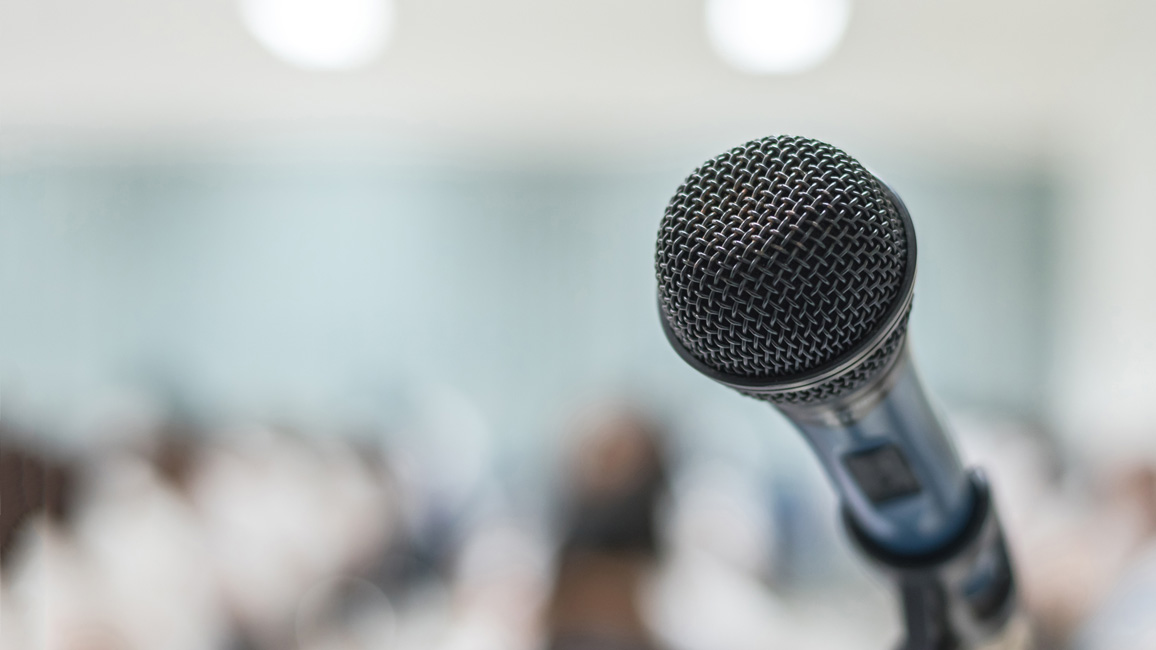 The picture shows a close-up of a table microphone in front of a blurred conference room.
