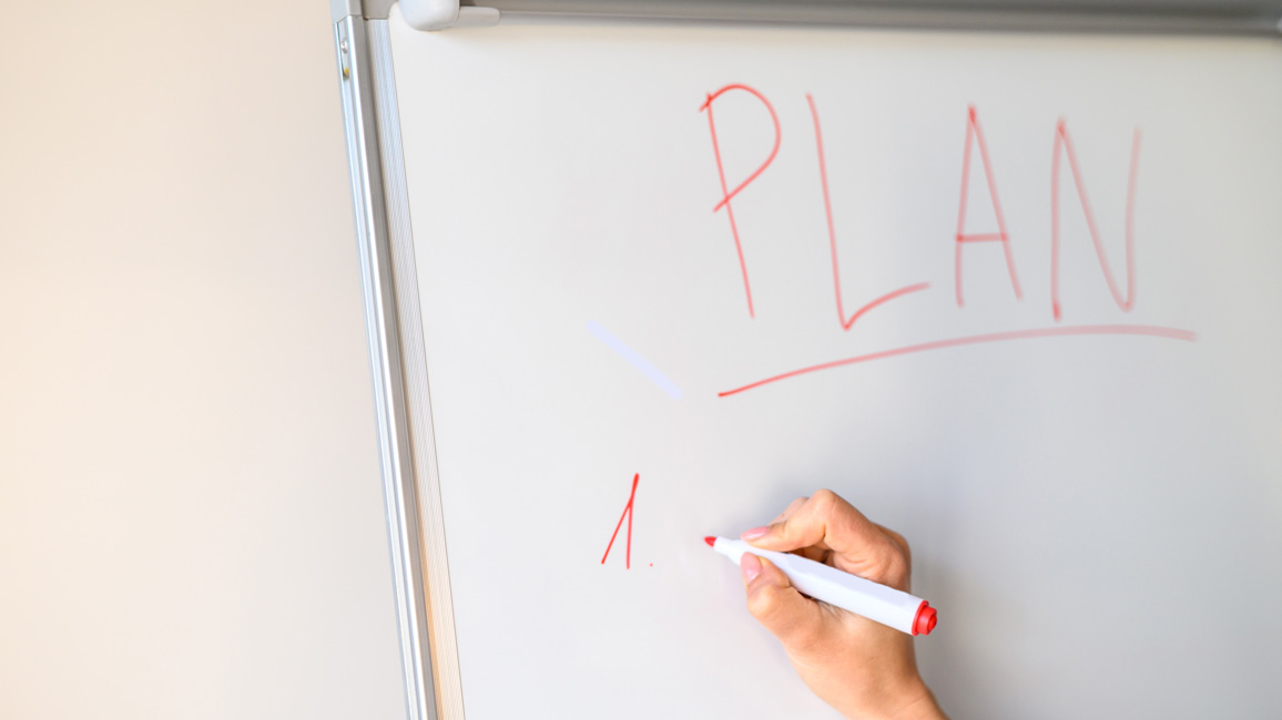 Eine Hand hält einen Stift und schreibt auf ein Whiteboard. Dort steht als Überschrift das Wort „Plan“ und darunter beginnt mit „1.“ eine Aufzählung.