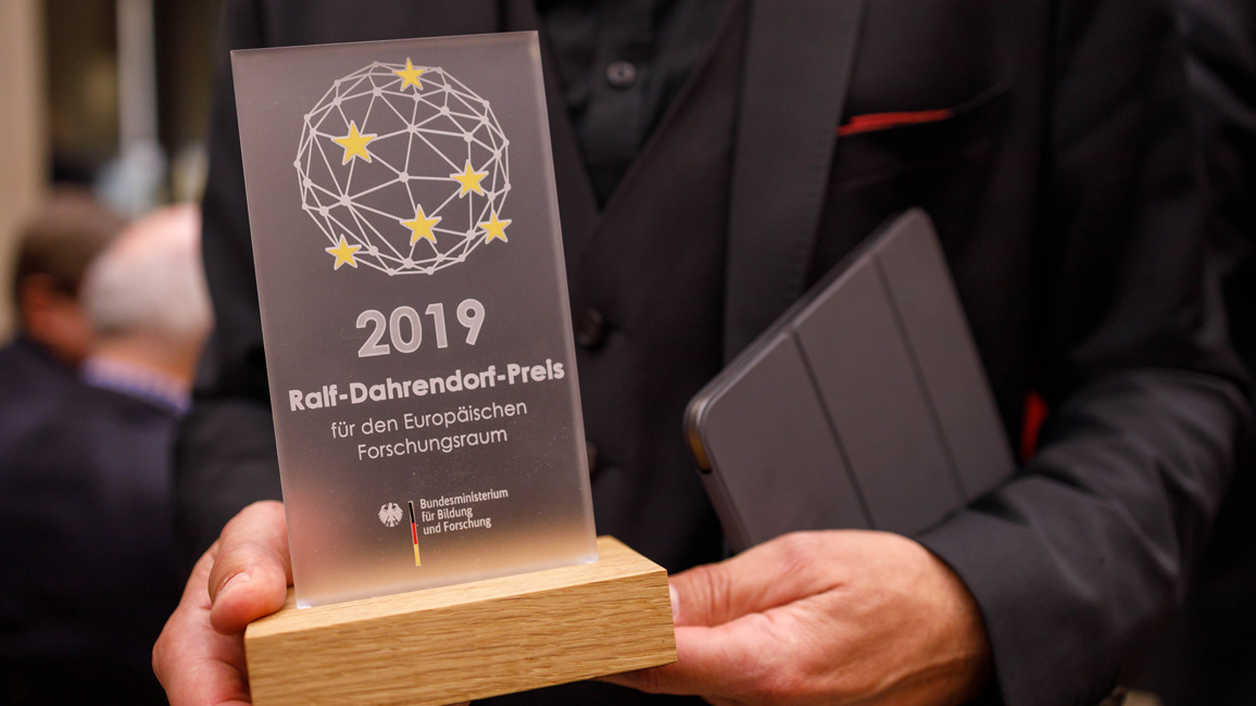 Ein Preisträger hält den Ralf-Dahrendorf-Preis für den Europäischen Forschungsraum in der Hand, unter seinem Arm ist eine Urkunde geklemmt. Der Preis ist eine Acrylscheibe mit Gravur, die auf einer quadratischen Holzscheibe befestigt ist.
