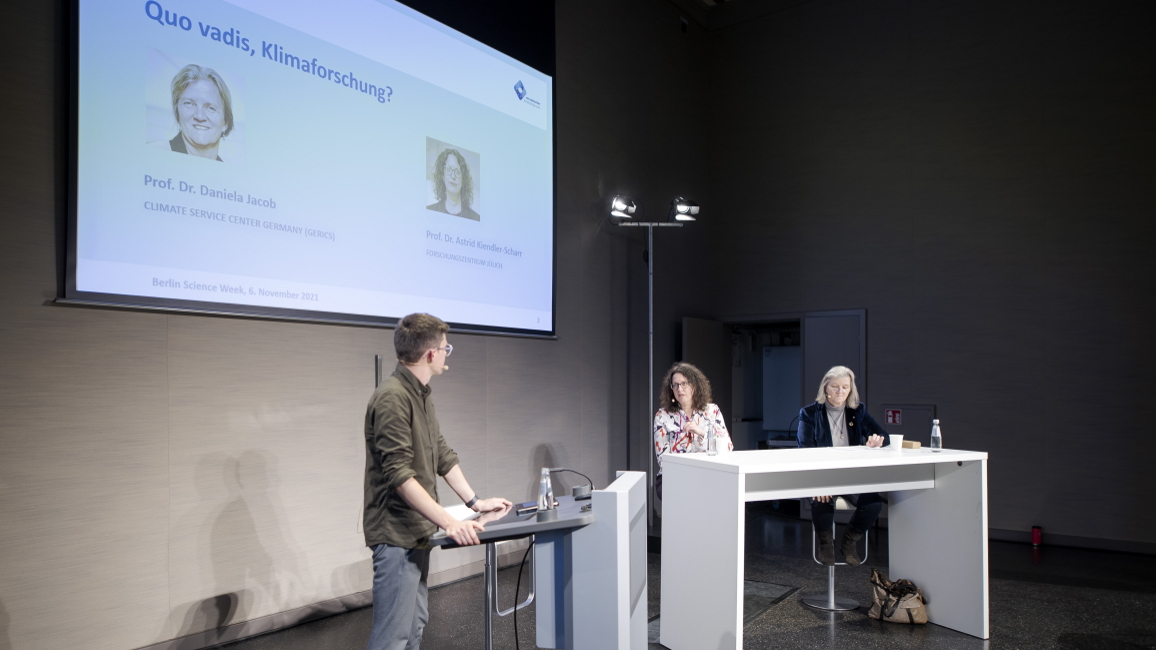 Panel-Diskussion auf der Bühne mit 2 weiblichen Panelistinnen und dem Moderator, der am Pult steht. Im Hintergrund ein großer Bildschirm.