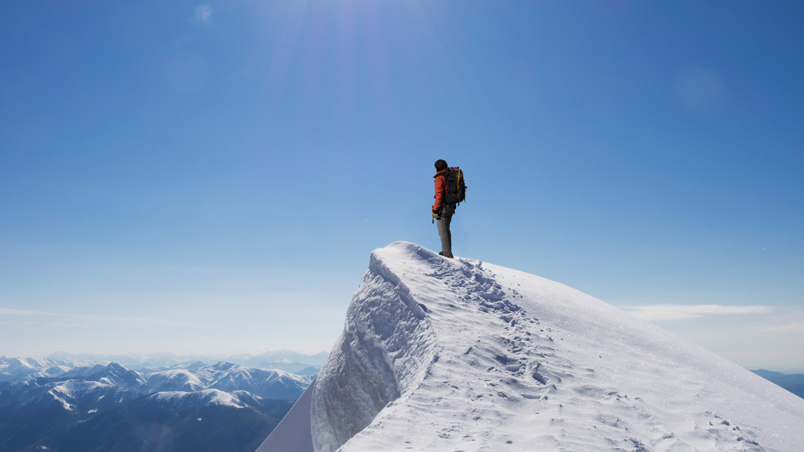 A mountain climber stands on the top of a mountain looking into the distance in front oft he blue sky.