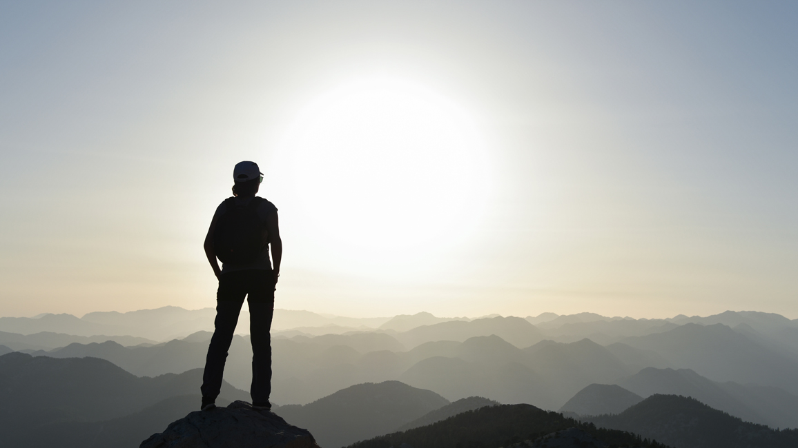 Eine nur als Silhouette zu sehende Person steht auf einem Berggipfel und sieht bei Sonnenaufgang in die Ferne.