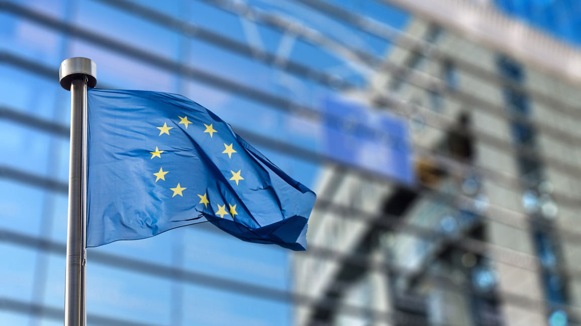 Eine EU-Flagge flattert an einem Fahnenmast vor dem Berlaymont-Gebäude in Brüssel, dem Sitz der Europäischen Kommission. Das Gebäude ist verschwommen ausschnittsweise im Hintergrund zu sehen.