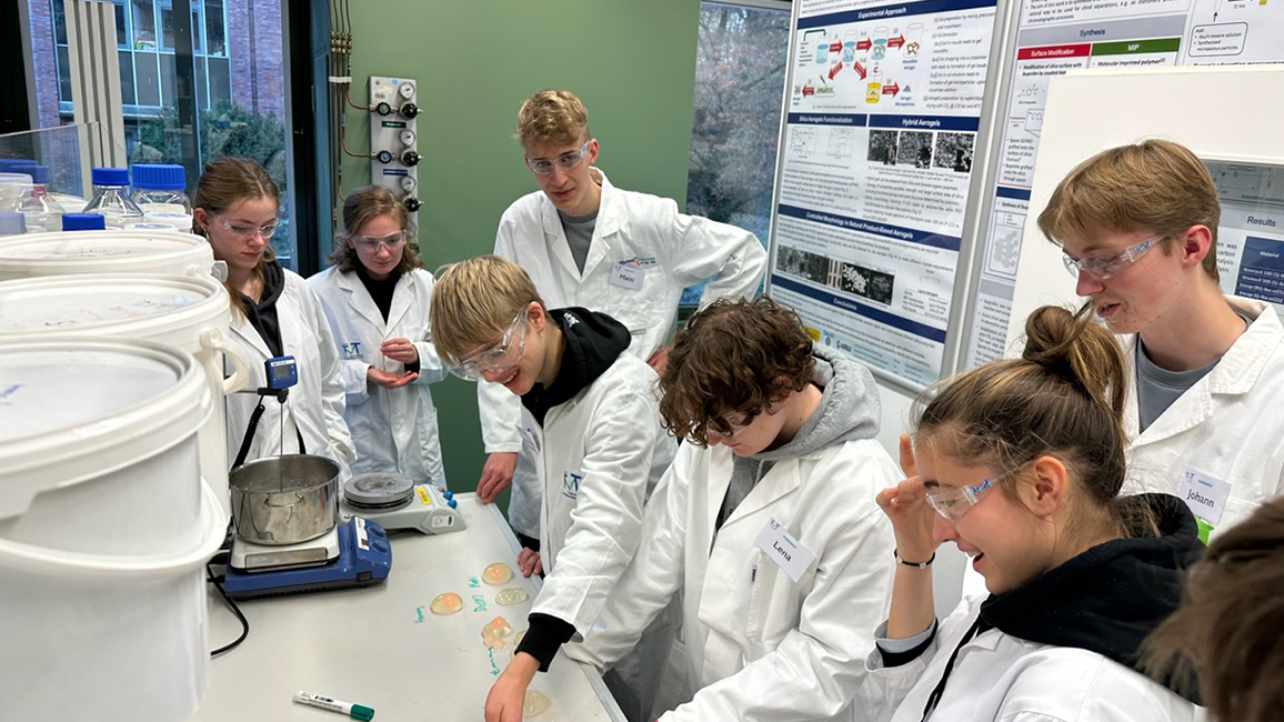 Schülerinnen und Schüler mit Schutzbrillen beugen sich über einen Tisch in einem Labor und arbeiten an einem Projekt. Im Hintergrund sind Info-Poster zu sehen.