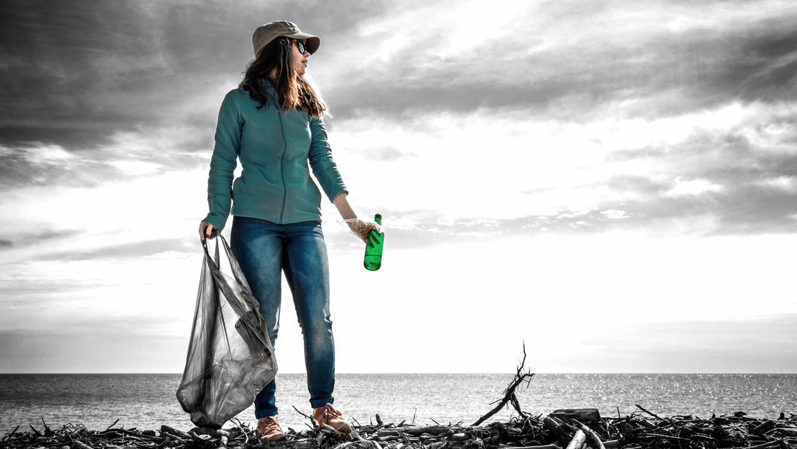 Eine Frau steht an der Küste und sammelt Flaschen in einem Müllbeutel. Im Hintergrund ist Wasser zu sehen.