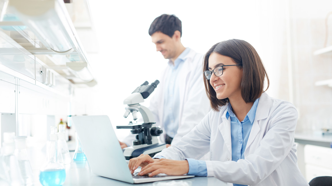 Eine Frau im Laborkittel tippt in ein Laptop, das vor einigen, mit bläulicher Flüssigkeit gefüllten Bechergläsern liegt. Im Hintergrund steht ein Mann im Laborkittel neben einem Mikroskop.